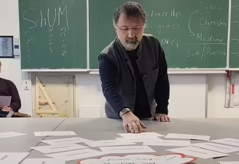 Referent an einem Tisch mit Kartenmaterial vor einer beschriebenen Tafel