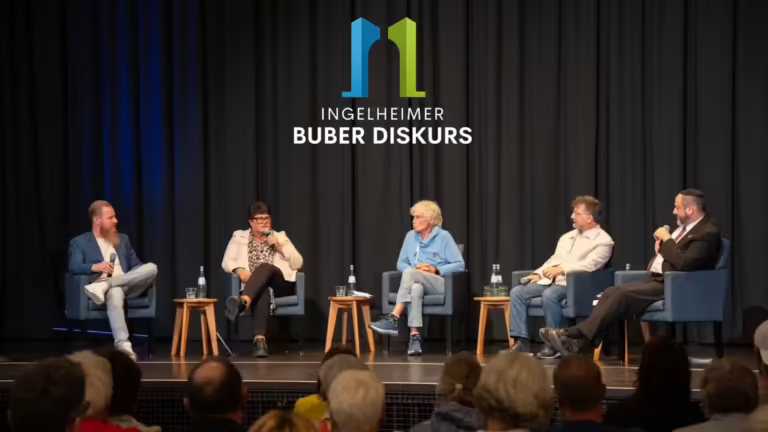 Das Panel des Ingelheimer Buber Diskurses auf der Bühne im WBZ Ingelheim vor Publikum, darüber das Logo des Ingelheimer Buber Diskurses